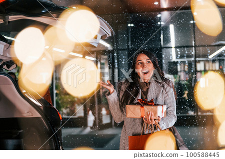 Near the car with gift box. Cheerful woman is outdoors at Christmas holidays time. Conception of new year Near the car with gift box. Cheerful woman is outdoors at Christmas holidays time. Conception of new year 100588445