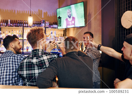 Cheering by knocking beer glasses. Group of people together indoors in the pub have fun at weekend time Cheering by knocking beer glasses. Group of people together indoors in the pub have fun at weekend time 100589231