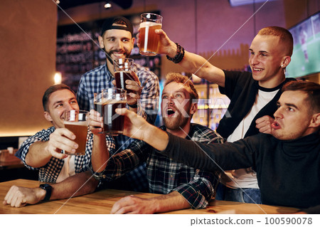 Cheering by knocking beer glasses. Group of people together indoors in the pub have fun at weekend time Cheering by knocking beer glasses. Group of people together indoors in the pub have fun at weekend time 100590078
