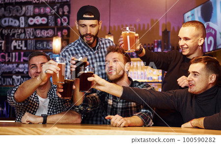 Cheering by knocking beer glasses. Group of people together indoors in the pub have fun at weekend time 100590282