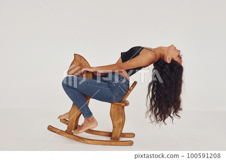Sits on kid's wooden horse toy. Young woman in jeans and bra indoors. White background 100591208