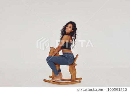 Sits on kid's wooden horse toy. Young woman in jeans and bra indoors. White background Sits on kid's wooden horse toy. Young woman in jeans and bra indoors. White background 100591210
