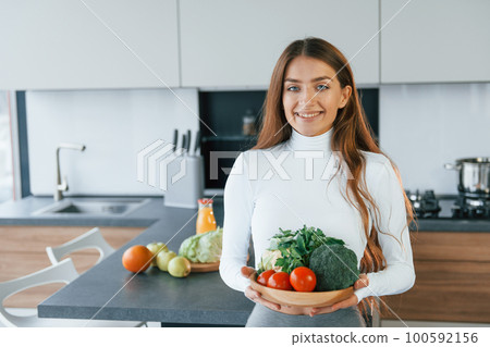 Holds plate with vegetables. Young european woman is indoors at kitchen indoors with healthy food Holds plate with vegetables. Young european woman is indoors at kitchen indoors with healthy food 100592156