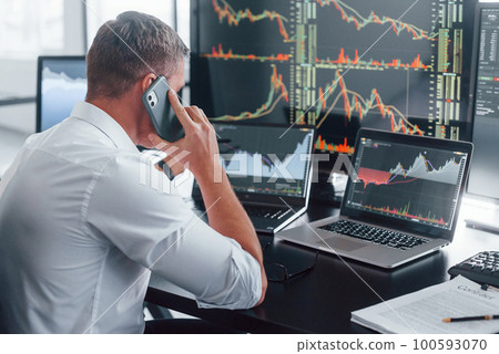 Talks by phone. Young male stock brocker works indoors in the office. Many of displays 100593070