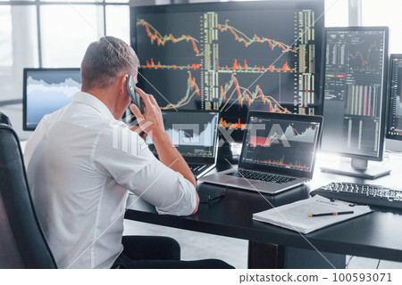 Talks by phone. Young male stock brocker works indoors in the office. Many of displays 100593071