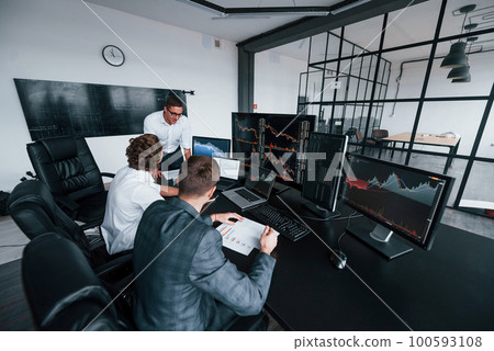 Holds document in hands. Team of stockbrokers works in modern office with many display screens 100593108