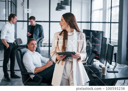 Young woman in formal wear standing of front of people. Team of stockbrokers works in modern office with many display screens Young woman in formal wear standing of front of people. Team of stockbrokers works in modern office with many display screens 100593236