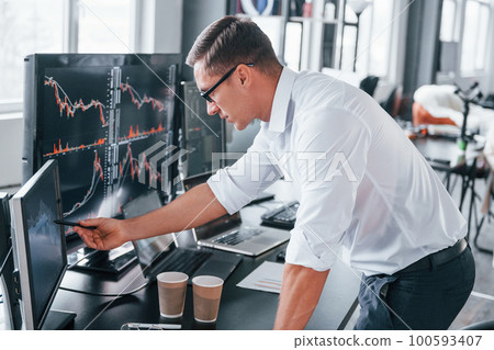 In formal clothes. Young male stock brocker works indoors in the office. Many of displays In formal clothes. Young male stock brocker works indoors in the office. Many of displays 100593407