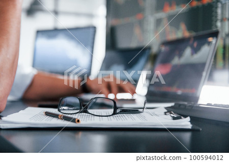 Close up focused view of glasses, notepad, documents and man at background Close up focused view of glasses, notepad, documents and man at background 100594012