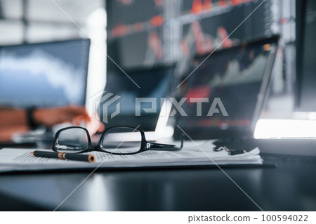 Close up focused view of glasses, notepad, documents and man at background Close up focused view of glasses, notepad, documents and man at background 100594022