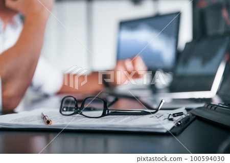 Close up focused view of glasses, notepad, documents and man at background Close up focused view of glasses, notepad, documents and man at background 100594030
