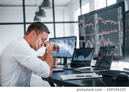 In formal clothes. Young male stock brocker works indoors in the office. Many of displays In formal clothes. Young male stock brocker works indoors in the office. Many of displays 100594105