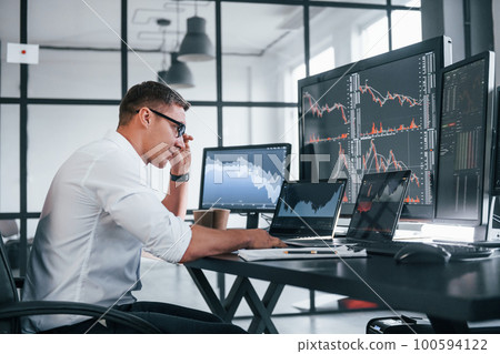 In formal clothes. Young male stock brocker works indoors in the office. Many of displays In formal clothes. Young male stock brocker works indoors in the office. Many of displays 100594122