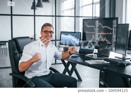 In formal clothes. Young male stock brocker works indoors in the office. Many of displays In formal clothes. Young male stock brocker works indoors in the office. Many of displays 100594125
