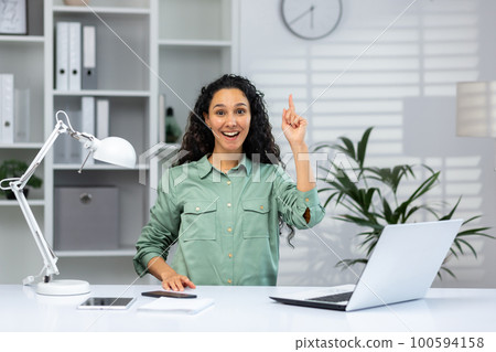 A young muslim woman sits in the office at a desk with a laptop, looks at the camera. points his finger up, indicates, has an idea, a plan. A young muslim woman sits in the office at a desk with a laptop, looks at the camera. points his finger up, indicates, has an idea, a plan. 100594158