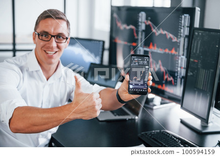 Holds phone. Young male stock brocker works indoors in the office. Many of displays Holds phone. Young male stock brocker works indoors in the office. Many of displays 100594159