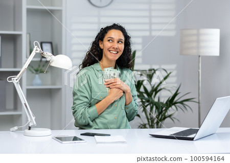 Young hispanic business woman sitting in office at desk and hugging money cash, looking satisfied at camera. He dreams of shopping, thinks about losses, purchases, investments. Young hispanic business woman sitting in office at desk and hugging money cash, looking satisfied at camera. He dreams of shopping, thinks about losses, purchases, investments. 100594164