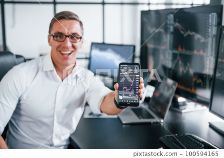 Holds phone. Young male stock brocker works indoors in the office. Many of displays Holds phone. Young male stock brocker works indoors in the office. Many of displays 100594165