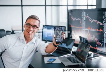 Holds phone. Young male stock brocker works indoors in the office. Many of displays Holds phone. Young male stock brocker works indoors in the office. Many of displays 100594170