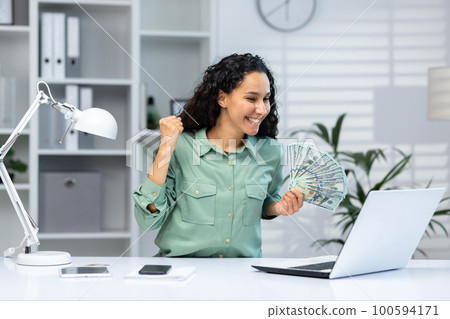 Online casino. A young Muslim woman sits in the office at a table with a laptop, holds cash money in her hand, plays online games, places bets, celebrates winnings. 100594171