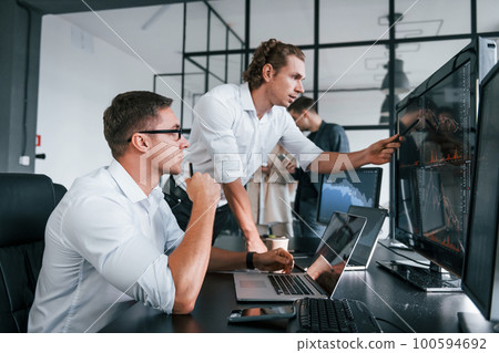 Sitting by the table. Team of stockbrokers works in modern office with many display screens Sitting by the table. Team of stockbrokers works in modern office with many display screens 100594692