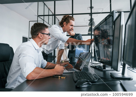 Guy in glasses concentrated at work. Team of stockbrokers is in modern office with many display screens Guy in glasses concentrated at work. Team of stockbrokers is in modern office with many display screens 100594699