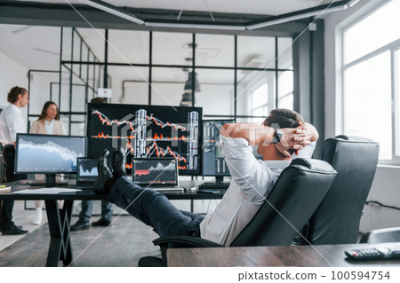 Man sits on chair and takes break. Team of stockbrokers works in modern office with many display screens 100594754