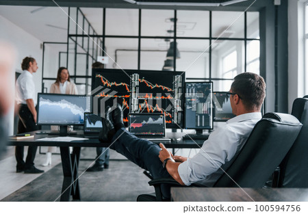 Man sits on chair and takes break. Team of stockbrokers works in modern office with many display screens Man sits on chair and takes break. Team of stockbrokers works in modern office with many display screens 100594756