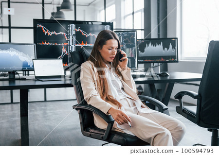 Talks by phone. Young female stock brocker works indoors in the office. Many of displays 100594793