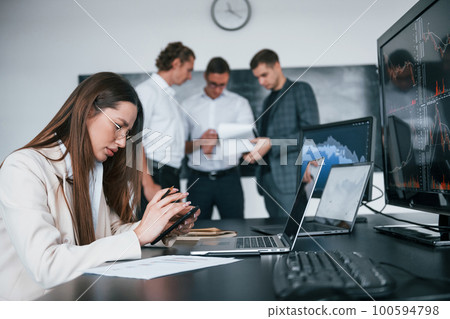 Woman sits in front of laptop and displays. Team of stockbrokers works in modern office with many display screens Woman sits in front of laptop and displays. Team of stockbrokers works in modern office with many display screens 100594798