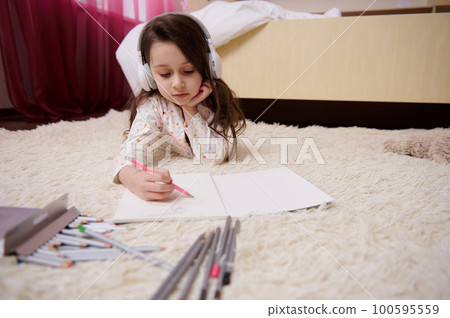 Caucasian talented little girl in pajamas, listening to music on wireless headphones, drawing picture with colorful pencils, lying down on carpet in her bedroom. Kids development and entertainment. Caucasian talented little girl in pajamas, listening to music on wireless headphones, drawing picture with colorful pencils, lying down on carpet in her bedroom. Kids development and entertainment. 100595559