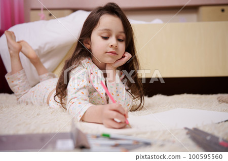 Caucasian little child, a baby girl in stylish pajamas with multicolored dots, drawing picture with colorful pencils indoors in her bedroom. Art. Creativity. Children. Lifestyles. Leisure. Hobby Caucasian little child, a baby girl in stylish pajamas with multicolored dots, drawing picture with colorful pencils indoors in her bedroom. Art. Creativity. Children. Lifestyles. Leisure. Hobby 100595560