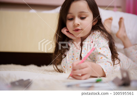 Adorable little girl with beautiful long hair, dressed in pajamas, lying down on the carpet in her pink bedroom, concentrated on drawing picture with colorful pencils. Kids. International Children's Adorable little girl with beautiful long hair, dressed in pajamas, lying down on the carpet in her pink bedroom, concentrated on drawing picture with colorful pencils. Kids. International Children's 100595561
