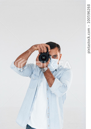 With professional camera. Young handsome man standing indoors against white background With professional camera. Young handsome man standing indoors against white background 100596268