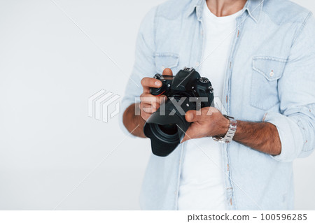 Holds camera in hands. Close up particle view of young man that standing indoors against white background Holds camera in hands. Close up particle view of young man that standing indoors against white background 100596285