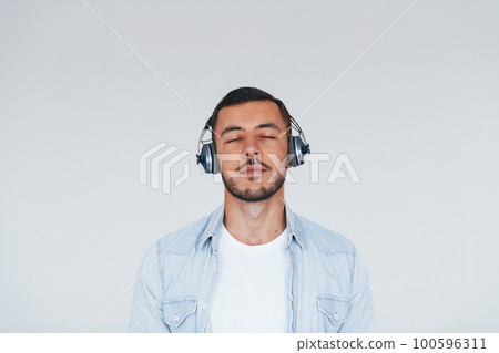 Listens to the music in headphones. Young handsome man standing indoors against white background 100596311