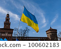 Flag with yellow and blue striped colors of Ukraine waving in the wind with a blue sky and sun. In the background the Castello Sforzesco in Milan. 100602183