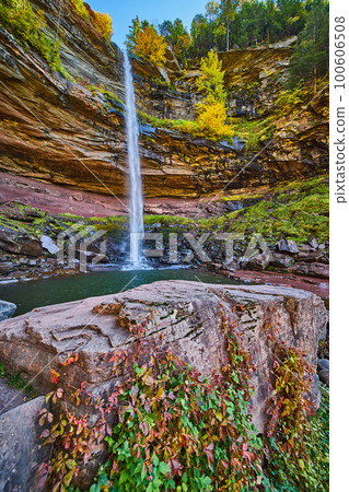 Waterfall pouring over layered cliffs from below with rocks with red and green vines climbing up 100606508