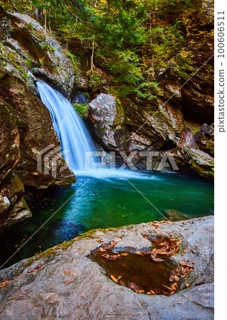 Puddle and leaves fill boulder by river of blue with waterfall over cliffs in forest Puddle and leaves fill boulder by river of blue with waterfall over cliffs in forest 100606511
