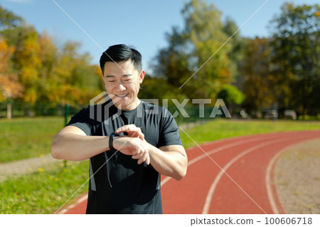 An Asian man, an athlete standing on a running track in a stadium, looking and checking the time on a smart watch. He smiles, satisfied with the result of the run, the marathon, breaking the record. 100606718