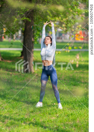 Young woman stretching body - warming up before running or working out. Female runner stretching arms before running at morning. Vertical photo. 100607408