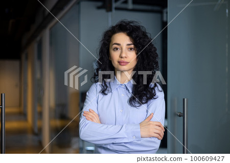 Portrait of serious female boss inside business company office, businesswoman crossed arms looking concentrated at camera, wearing shirt, satisfied and successful hispanic woman with curly hair. Portrait of serious female boss inside business company office, businesswoman crossed arms looking concentrated at camera, wearing shirt, satisfied and successful hispanic woman with curly hair. 100609427