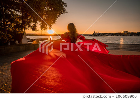 Sunrise red dress. A woman in a long red dress against the backdrop of sunrise, bright golden light of the sun's rays. The concept of femininity, harmony. 100609906