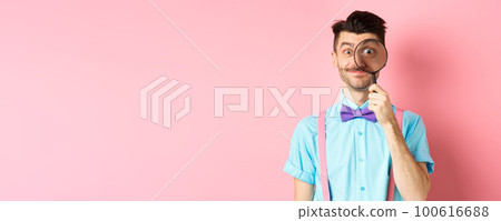 Funny caucasian man with moustache and bow-tie, looking through magnifying glass with big eye and smiling, standing over pink background 100616688