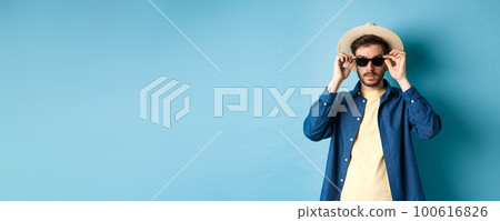 Handsome tourist in straw hat put on sunglasses on summer vacation, standing on blue background 100616826