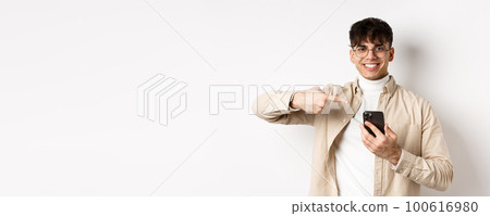 Real people. Handsome young man in glasses pointing finger at smartphone screen, showing online promo, standing on white background 100616980