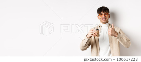 Real people. Cheerful smiling guy in glasses drinking coffee and showing thumb up, recommending cafe, standing on white background Real people. Cheerful smiling guy in glasses drinking coffee and showing thumb up, recommending cafe, standing on white background 100617228