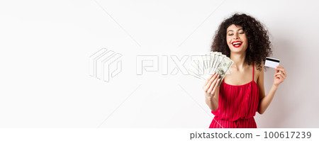 Shopping. Rich successful woman with curly hair and red dress, holding plastic credit card and looking pleased at dollar bills, white background Shopping. Rich successful woman with curly hair and red dress, holding plastic credit card and looking pleased at dollar bills, white background 100617239
