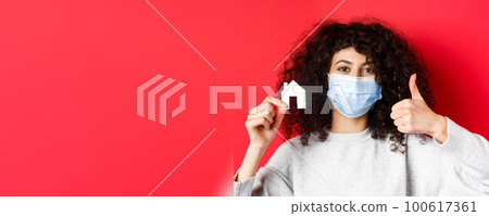 Real estate and pandemic concept. Close-up of woman recommending agency, wearing medical mask, showing thumbs up and paper house cutout, red background 100617361