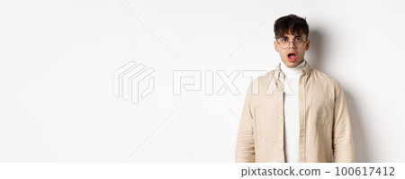 Portrait of shocked young man in glasses drop jaw, open mouth and stare amazed at camera, looking at something impressive, standing on white background 100617412
